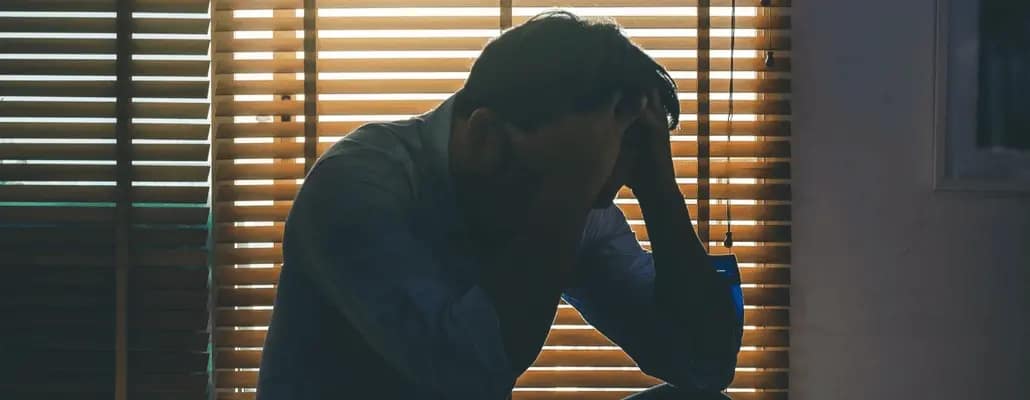 Homem com depressão sentado numa cama com cortina fechada.