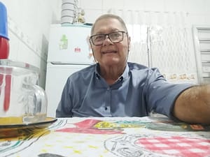 Homem com óculos e camisa branca sentado num banco de madeira, com sorriso no rosto.