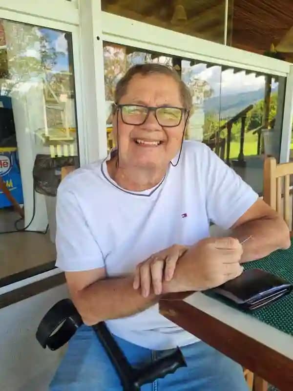 Homem com óculos e camisa branca sentado num banco de madeira, com sorriso no rosto.